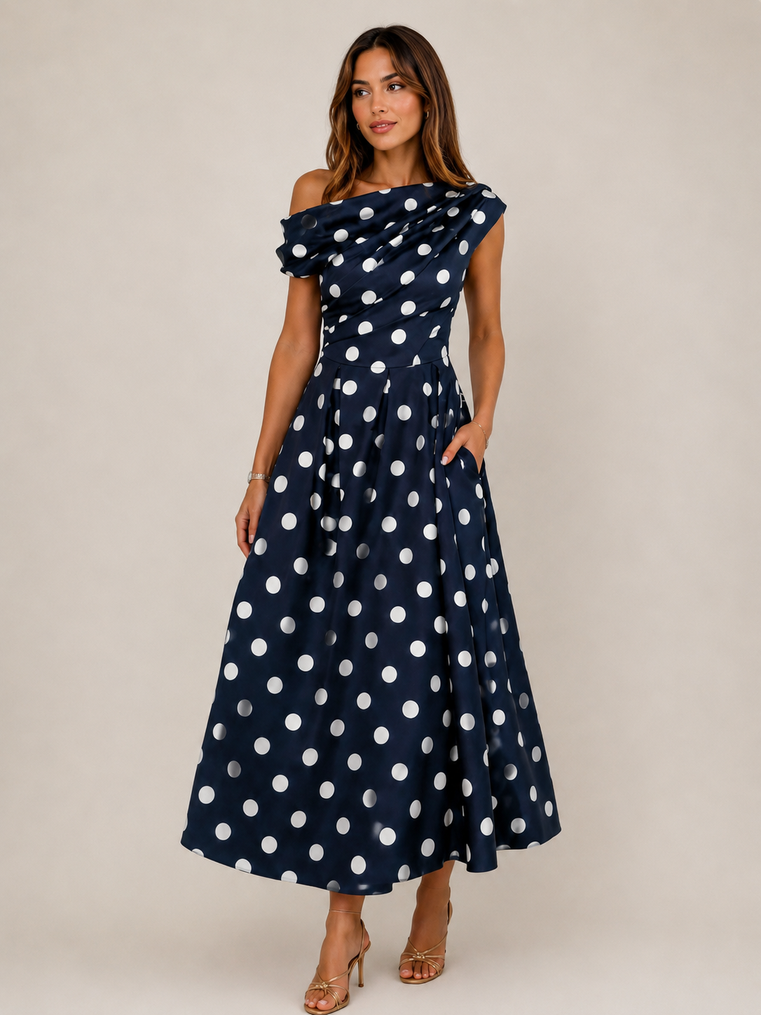 A woman modeling a navy blue off-the-shoulder maxi dress with white polka dots, standing against a neutral beige studio background.