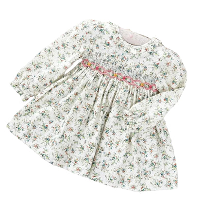 A white floral hand-smocked dress with ruffled collar and cuffs, and a sash tie at the back, displayed on a flat surface.