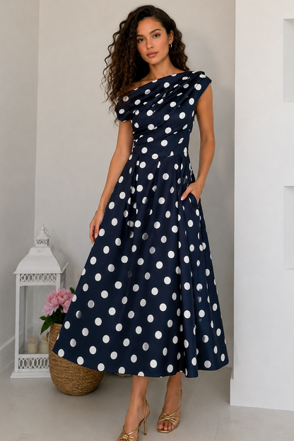 A woman models a navy blue midi dress with white polka dots, featuring an off-the-shoulder neckline, cap sleeves, and a flared skirt with pockets, set against a minimalist white interior background.